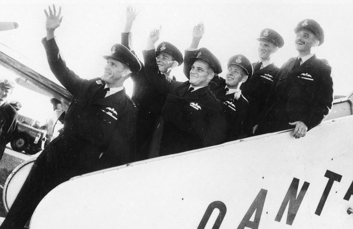 Six men in RAAF uniform smiling and waving while standing on the moveable staircase attached to a plane.