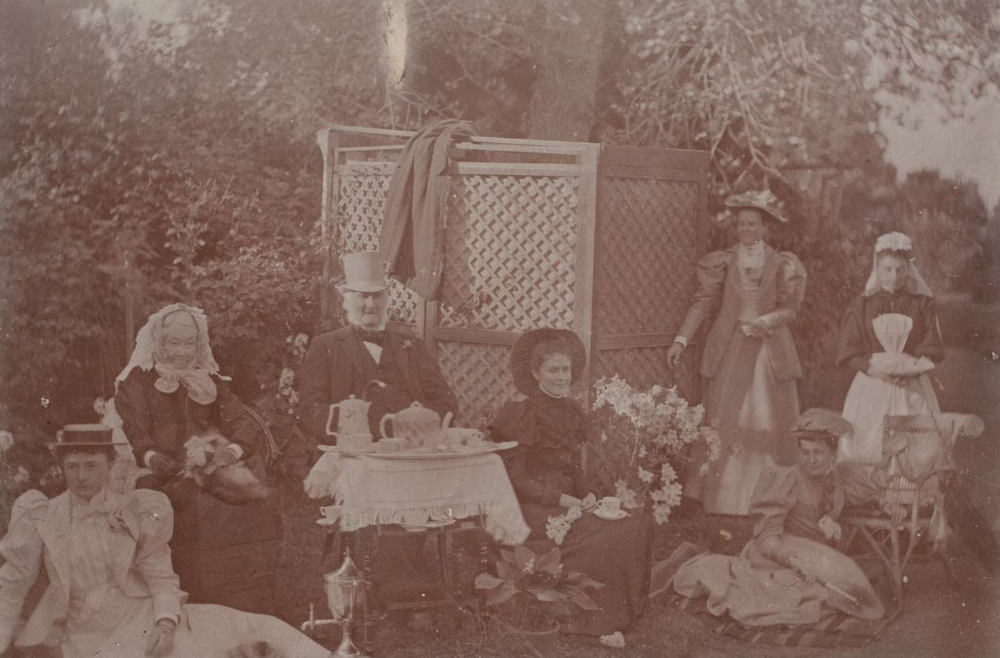 Sepia photograph showing a man and six women taking tea outside in a garden.