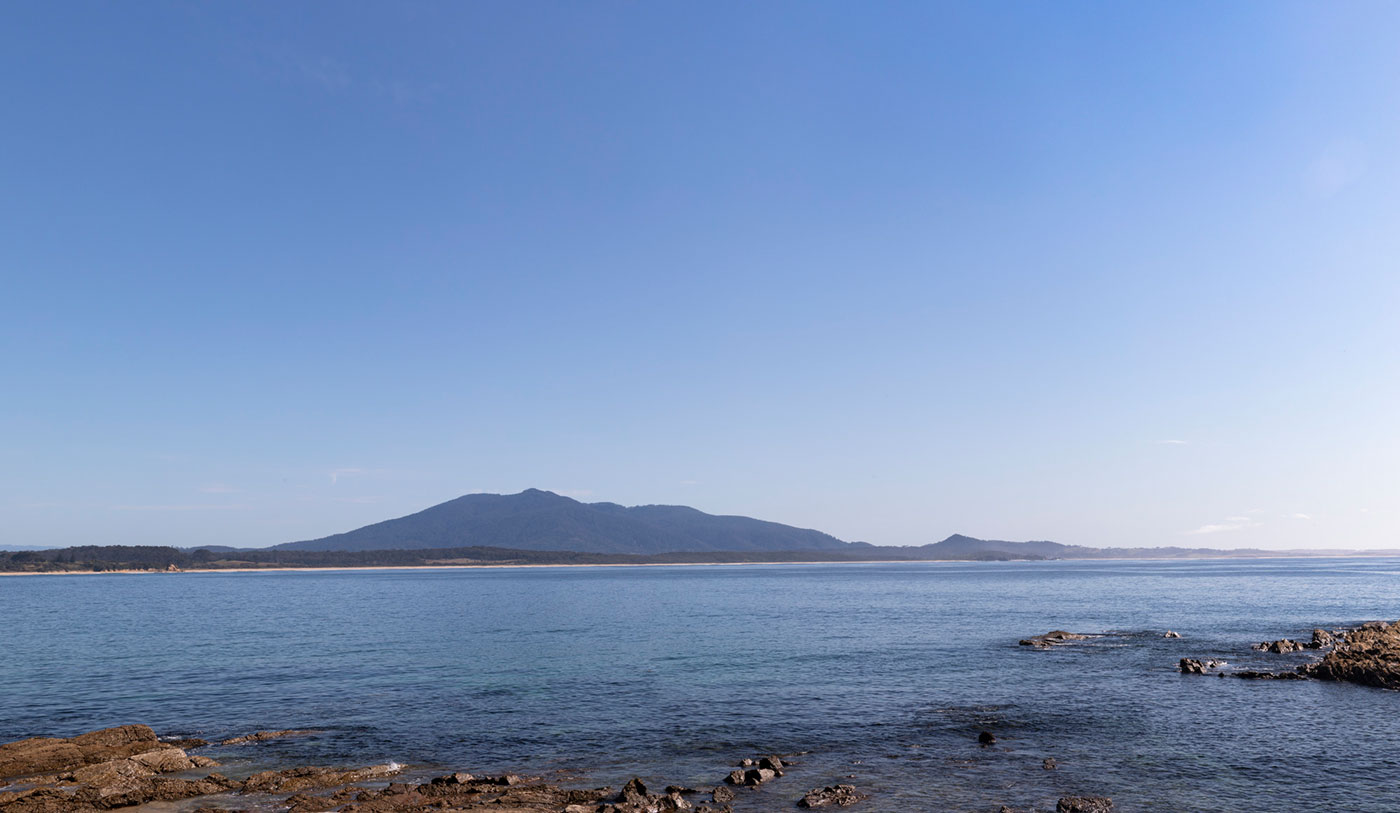 Colour photo of a bay with rocks in the foreground and a beach and mountain in the background.