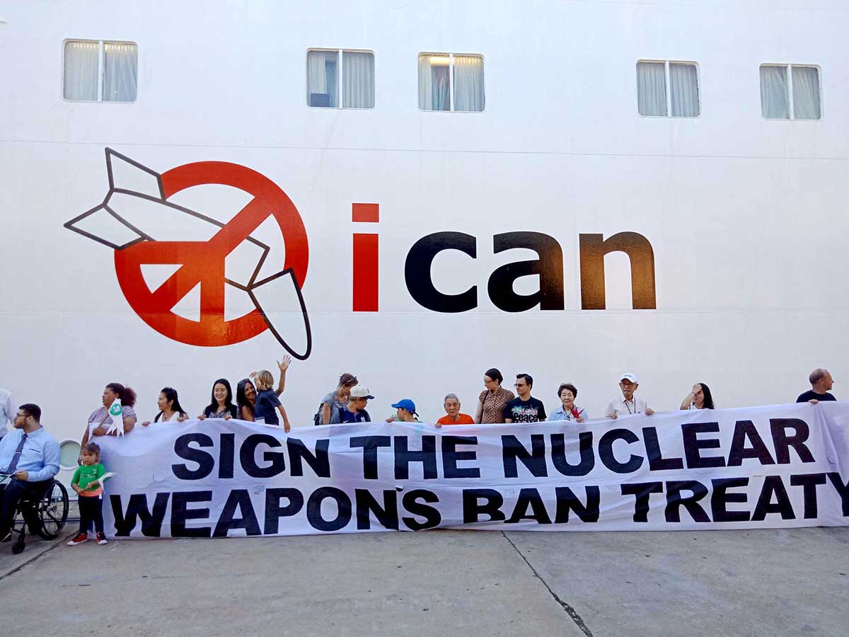 Colour photo of a group of people protesting and holding up a banner with the text ’SIGN THE NUCLEAR WEAPONS BAN TREATY’. They are standing in front of a large vessel or building with a logo painted onto its side. The logo features the abbreviation 'ican' and a red graphic featuring a peace sign overlapping a nuclear missile that has been broken in half.