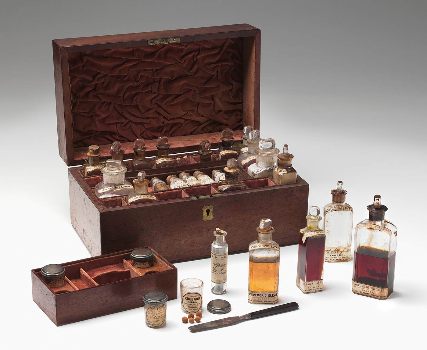 Wooden medicine chest with hinged lid. The interior is lined in red velvet with compartments around the edge and one central compartment holding two trays that lift out. The chest contains 27 bottles and jars holding medicinal substances and a small knife.