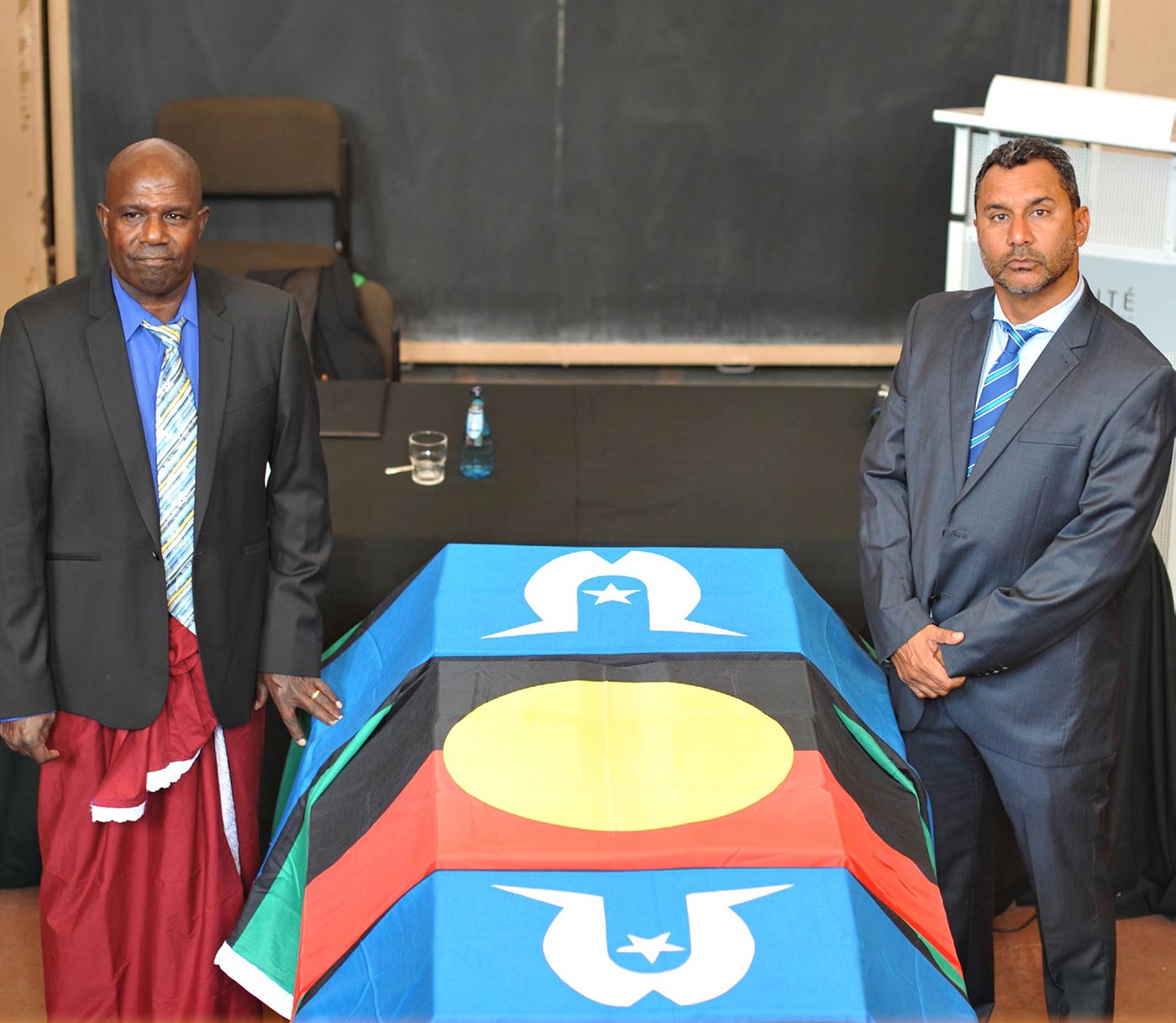 Photo of two men standing around a container covered by flags.