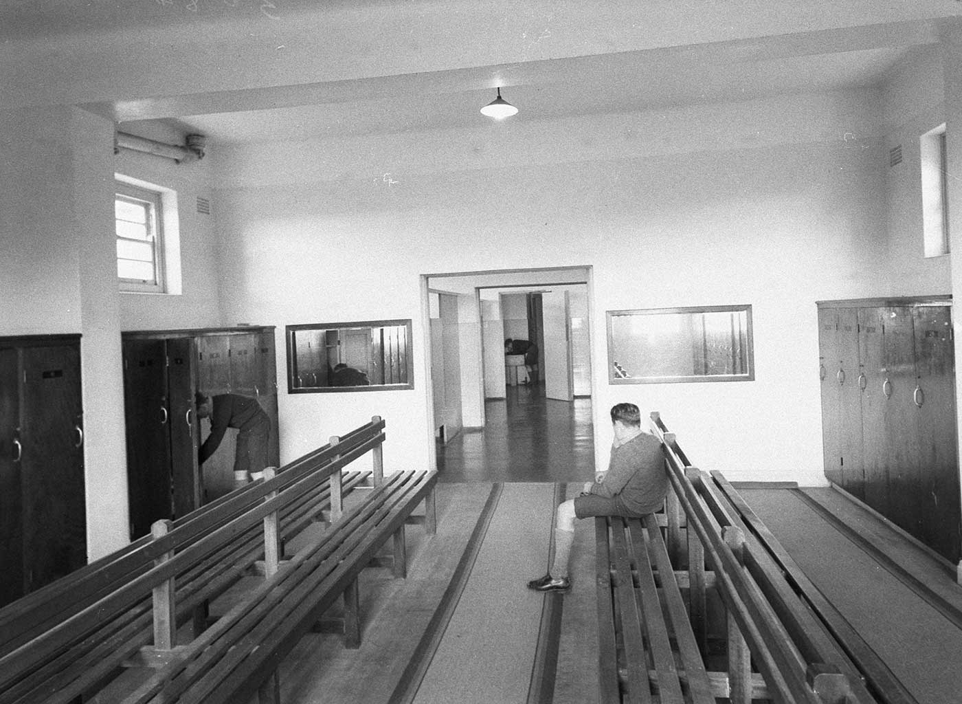 Black and white photo showing a room with wooden lockers lining the walls and two sets of long wooden benches at the centre. A boy in school uniform sits with his back to the camera, tying a shoelace, another boy leans into a locker and, at the far end of the room, a third boy leans over a sink.