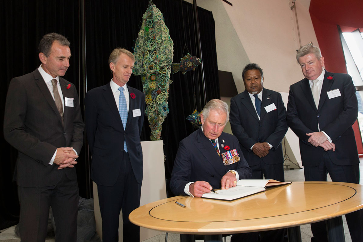 Prince Charles sits, signing a book. Behind him is a ghost net sculpture and four men, standing.
