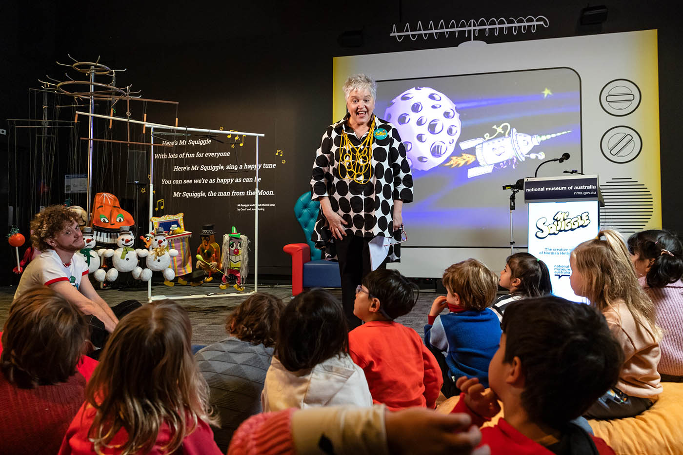 A woman stands before a large group of seated children, with an image of the moon and rocket projected on a screen behind.