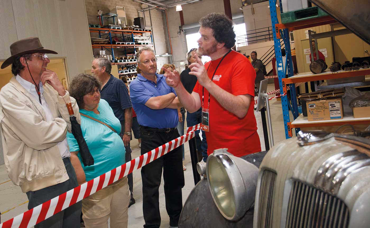 A photograph of three people looking on while a person standing behind the striped tape gives a talk.