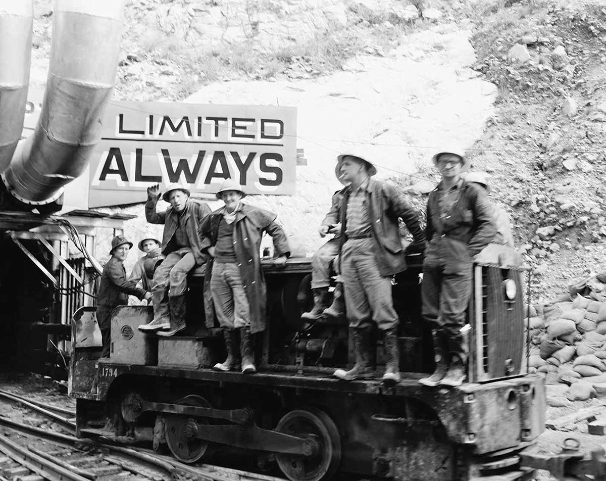 Photo of men on an open rail carriage outside a tunnel, with a large rock wall behind. the Snowy Hydro.