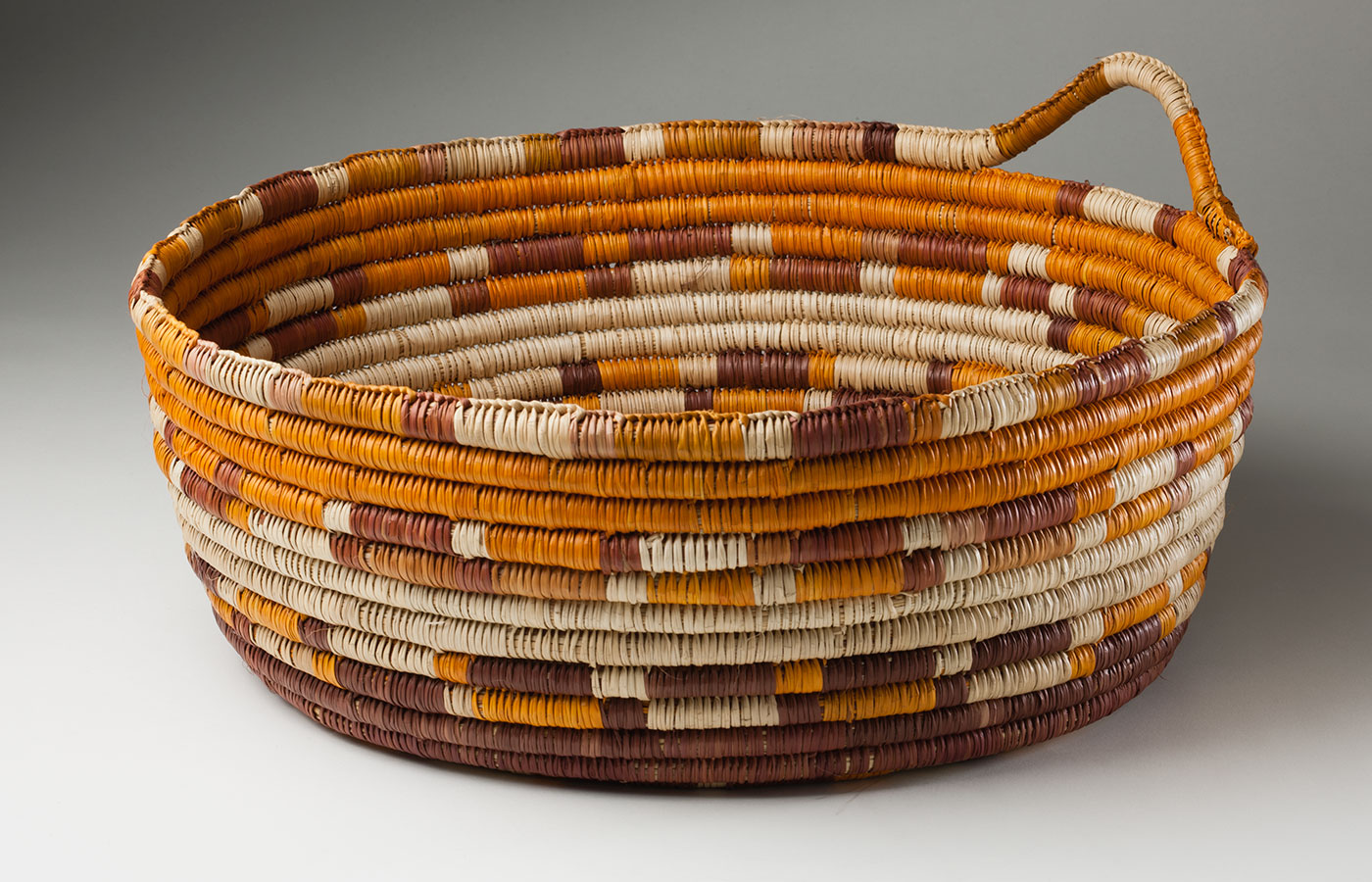 An orange, brown and straw-coloured coil basket made from pandanus fibre and vegetable dyes. It has an 'INJALAK' tag tied to a small handle at the side.