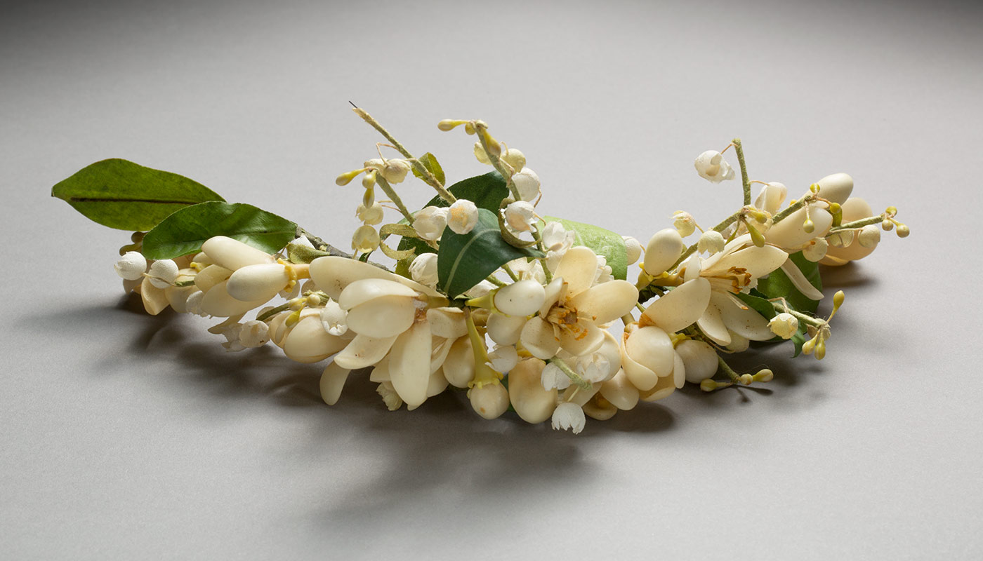 A strip of cream coloured fabric and wax or plastic flowers with green leaves. The flowers are on a curved piece of wire and are a little dirty.