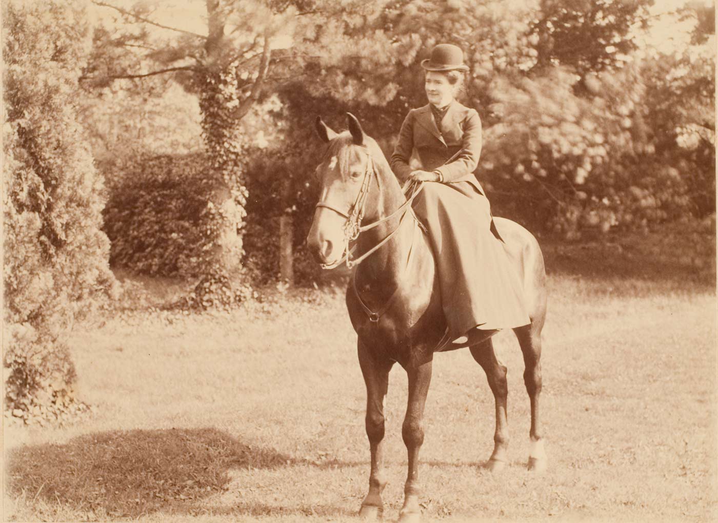 Sepia photo of a woman sitting side-saddle on a horse. She wears a long skirt, tailored jacket and hat.