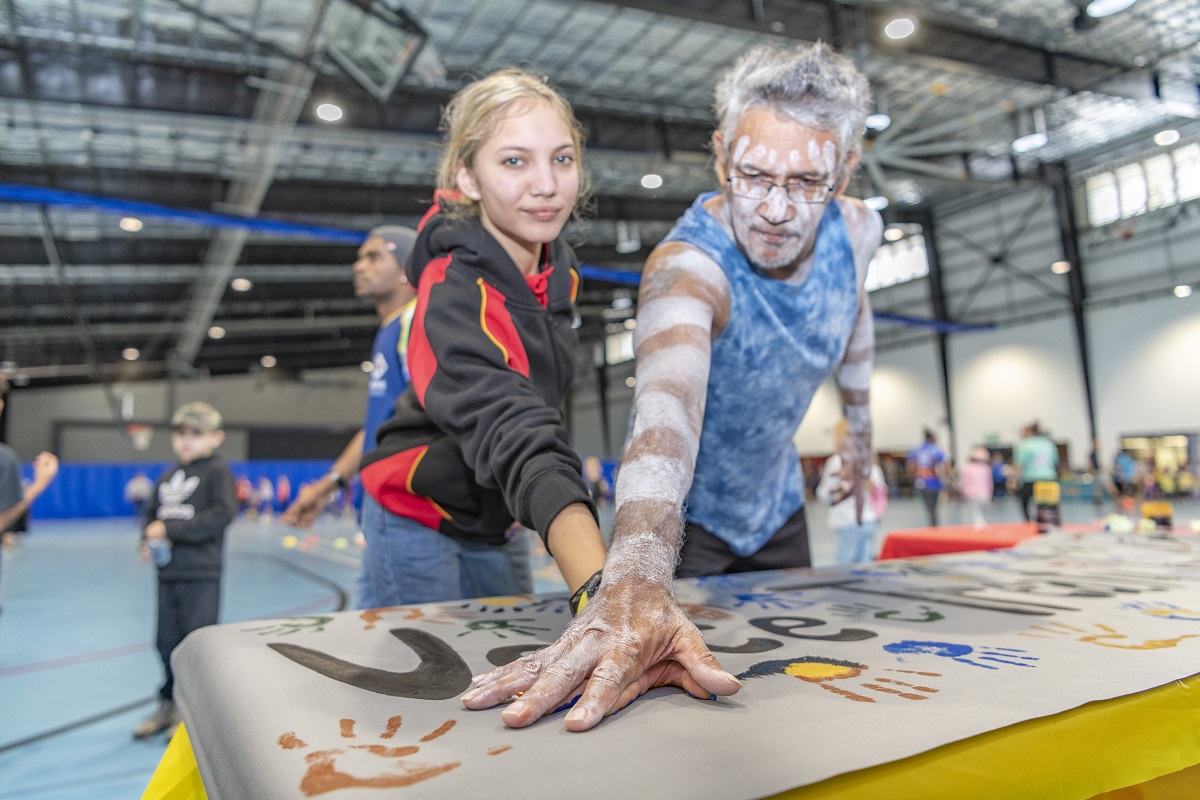 A photo of a young girl standing next to a man adorned with white body paint on his face and arms. They are placing their hands in paint and then onto paper for sign-making.