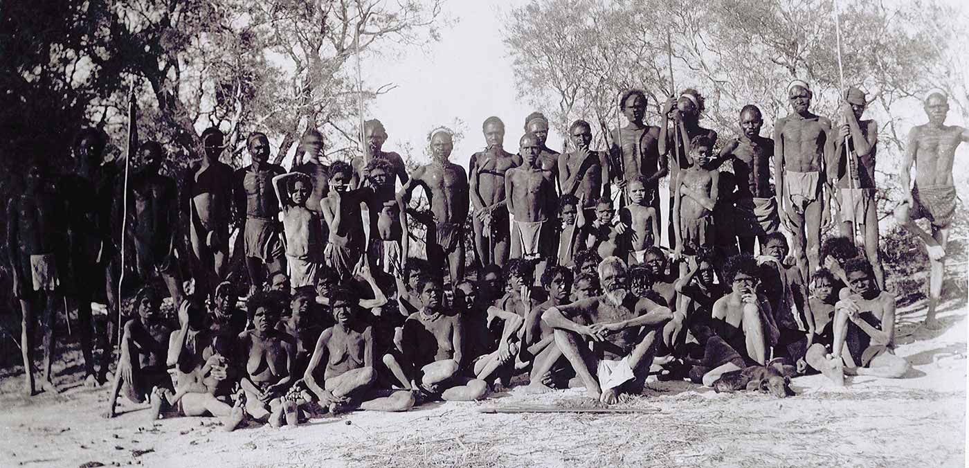 Black and white photo of a group photo of a large group of people in bushland. Some are holding spears.