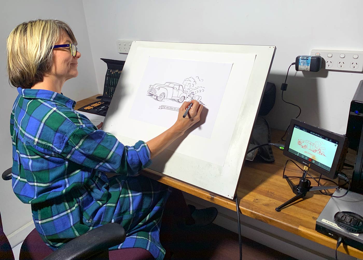 A woman sits at a studio desk sketching an animation using digital equipment.