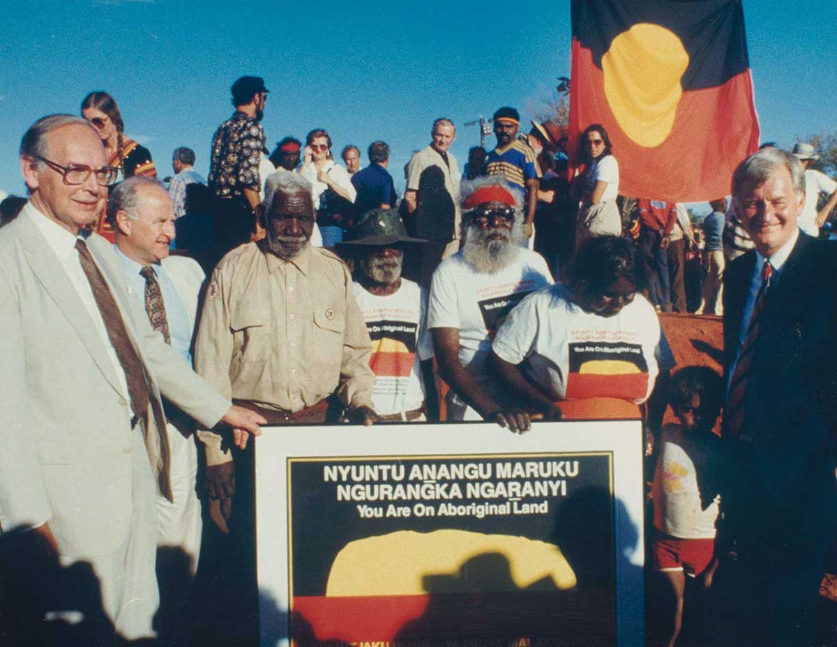 Colour photo of a crowd of people with the Aboriginal flag in the background. In the foreground are several people gathered around a large sign with the words