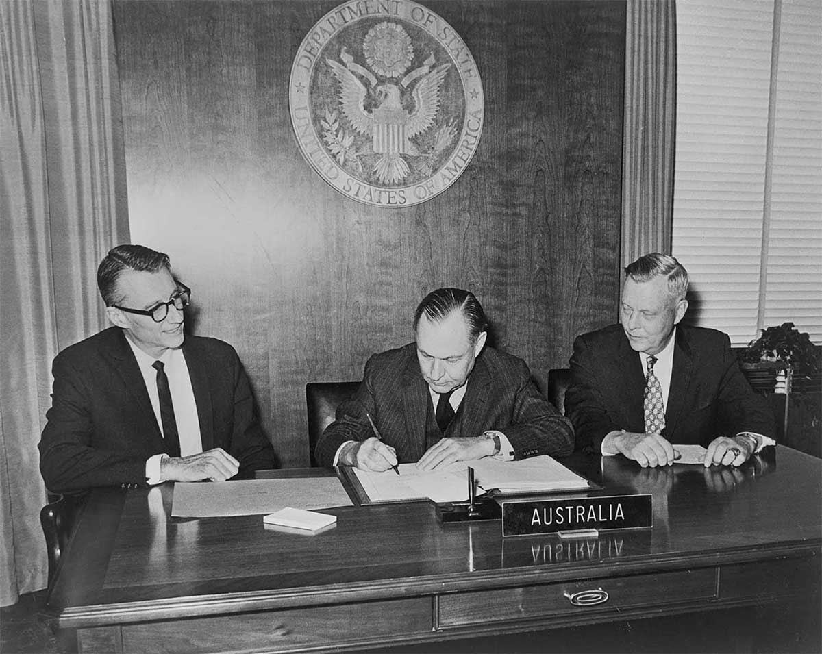 Black and white photo of three men sitting at a bureau and signing a document. There is a label sitting on the desk with the text ‘AUSTRALIA’ and there is an emblem mounted on the wall behind them with the text ‘DEPARTMENT OF STATE. UNITED STATES OF AMERICA’.