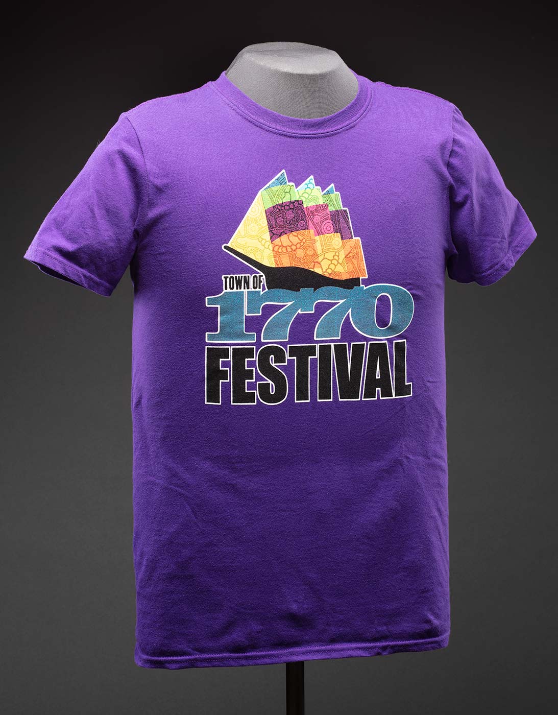 A purple T-shirt with a logo on the front that features a colourful, abstract design of a sailing ship with an indigenous design. Below the illustration is text that reads 'TOWN OF / 1770 / FESTIVAL'.