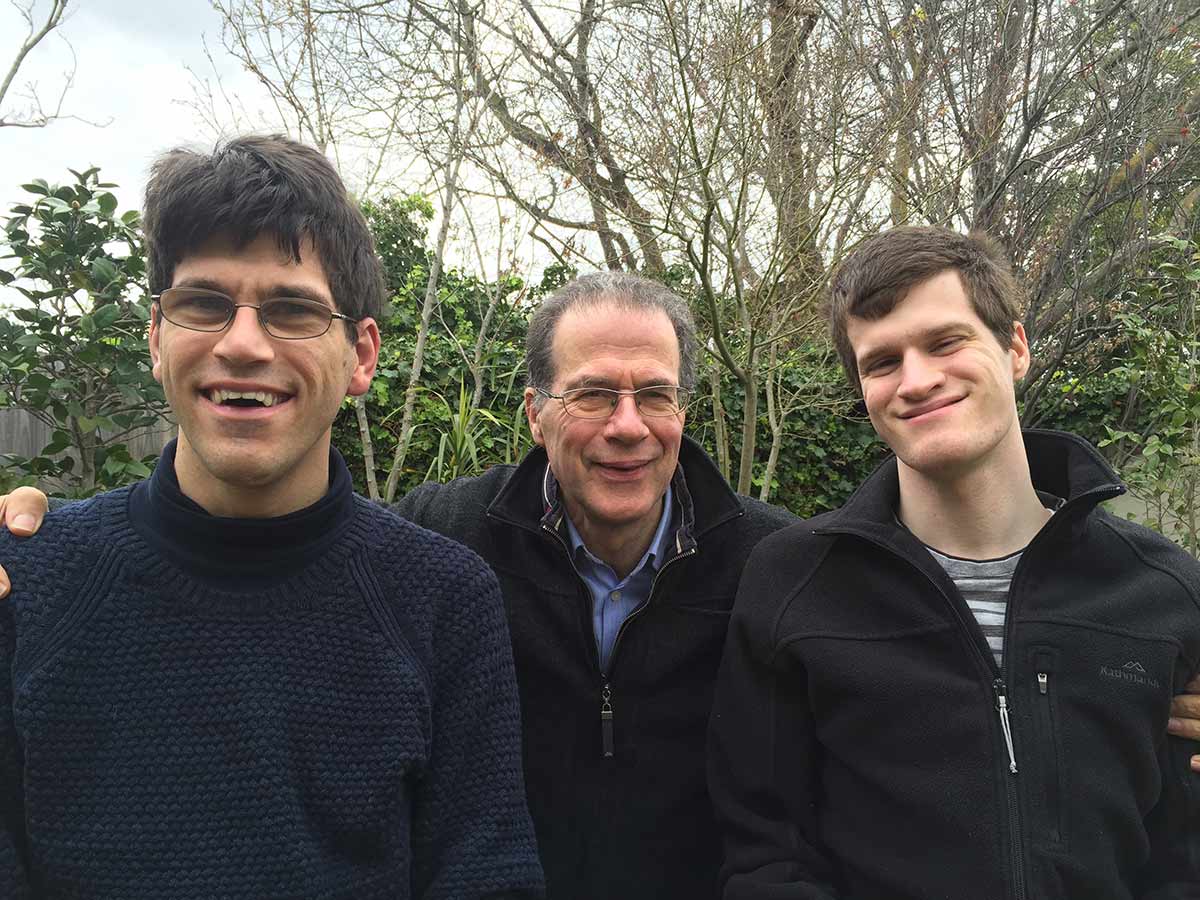 An older man and two younger men who are standing amongst a garden backdrop smile for the camera.