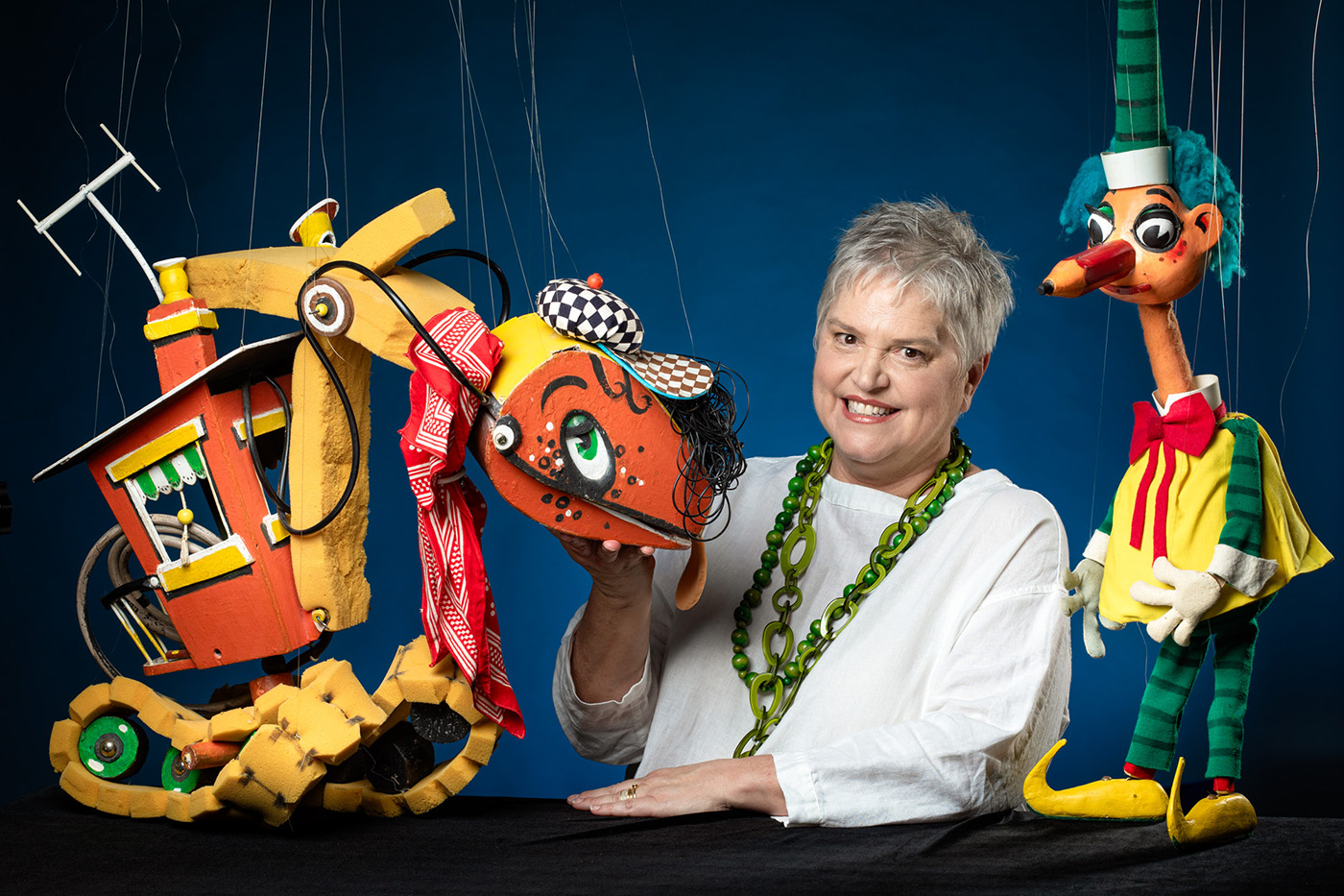 Photograph of Rebecca Hetherington with two characters. Rebecca has grey spikey hair and is wearing a white top and green necklace. On the left is Bill Steamshovel, an orange and yellow puppet of a steamshovel and to her right is Mr Squiggle.
