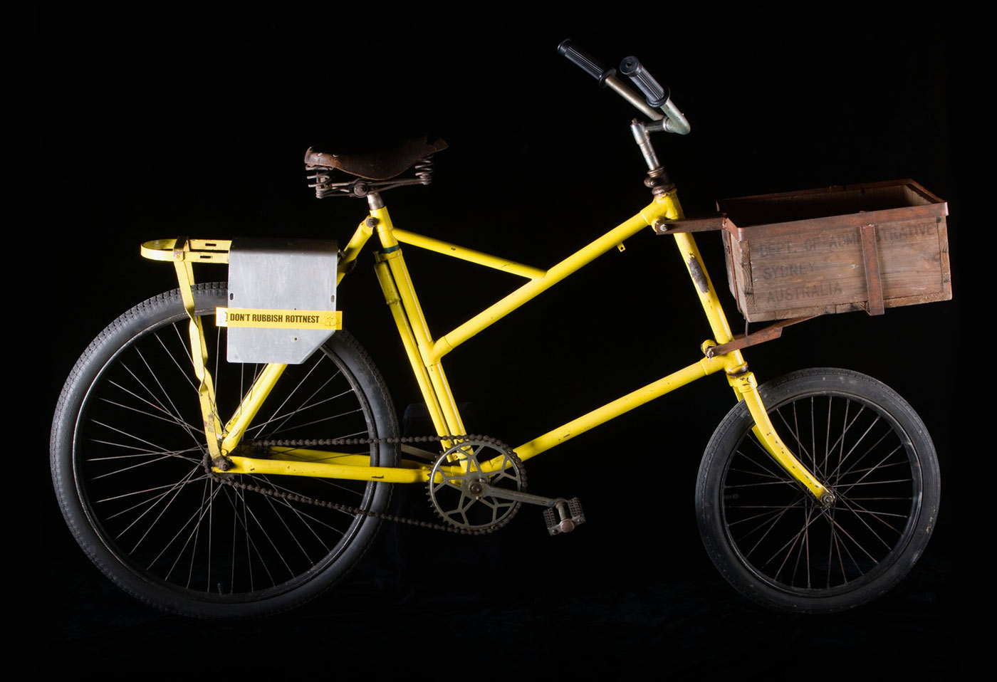 A yellow bicycle with a wooden crate on the front and a small detachable a yellow sticker on the back of the bicycle reads