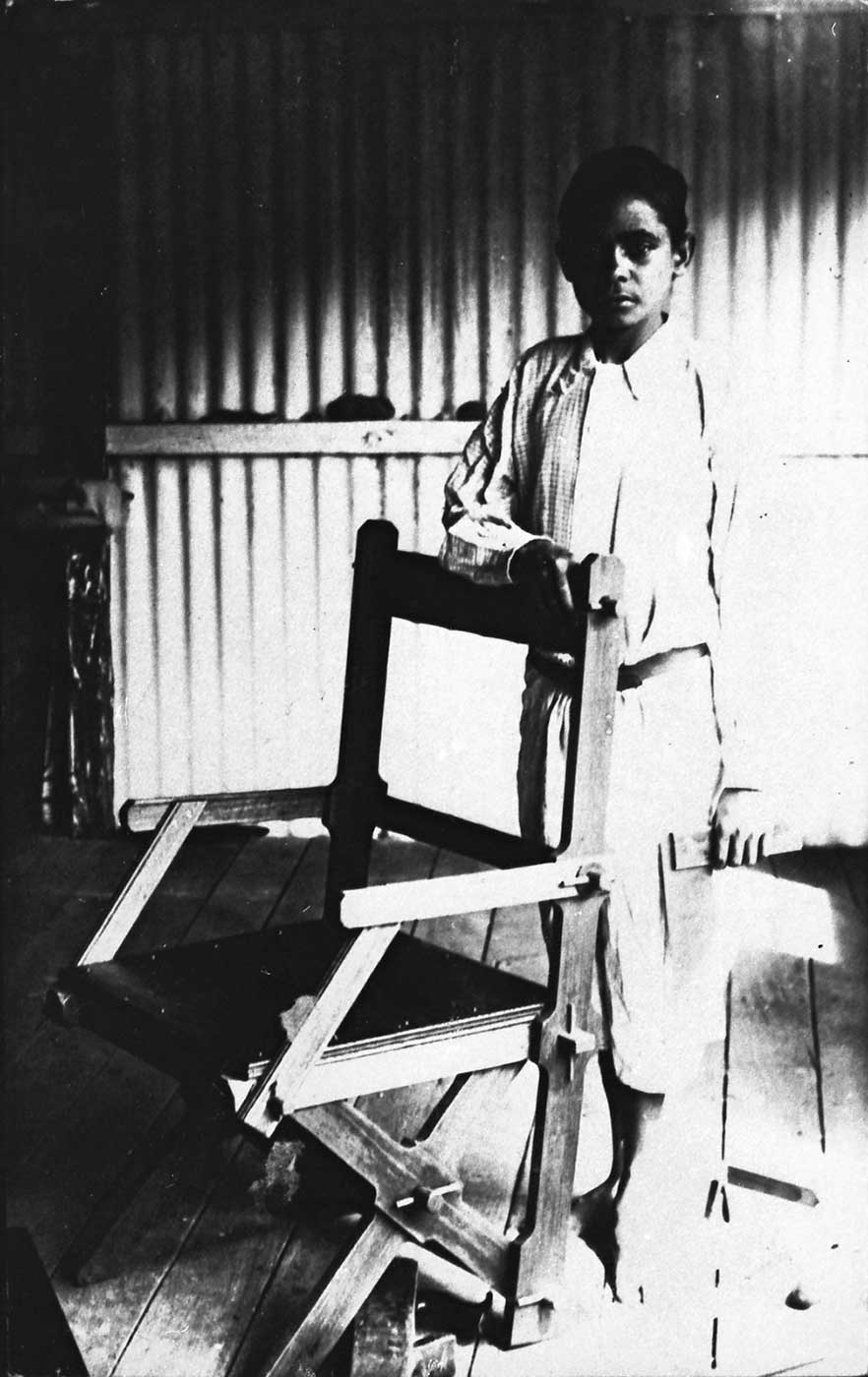 Black and white photograph of a boy standing behind a wooden chair. The boy is dressed in a shirt and short pants and is barefoot.