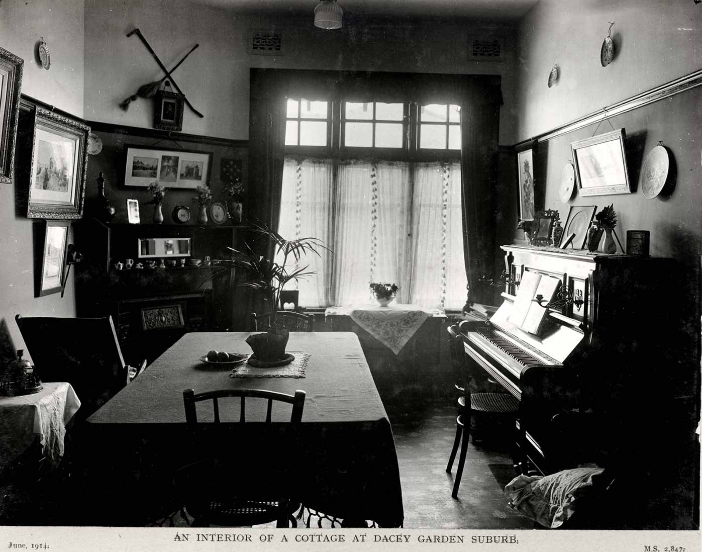 Black and white photograph showing part of a dining room, with a table at centre and piano at right.