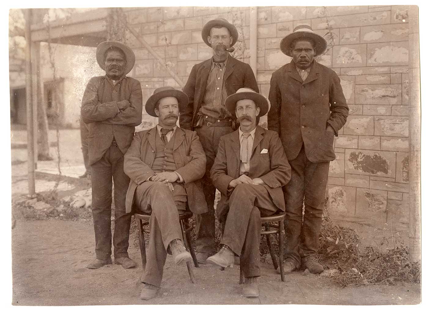 Sepia photograph showing two men seated, with their legs crossed. Three men stand behind them.