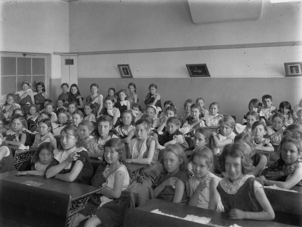 Black and white photograph showing a classroom of young girls, seated at desks. Many are lifting their left sleeve so show their upper arm.