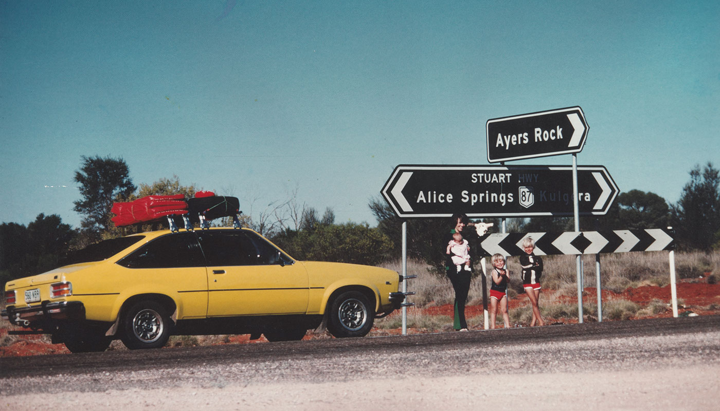 Colour photo showing a woman and three children in front of a road junction, with signs pointing to 'Ayers Rock' and 'Alice Springs'. Beside them is a bright yellow car.