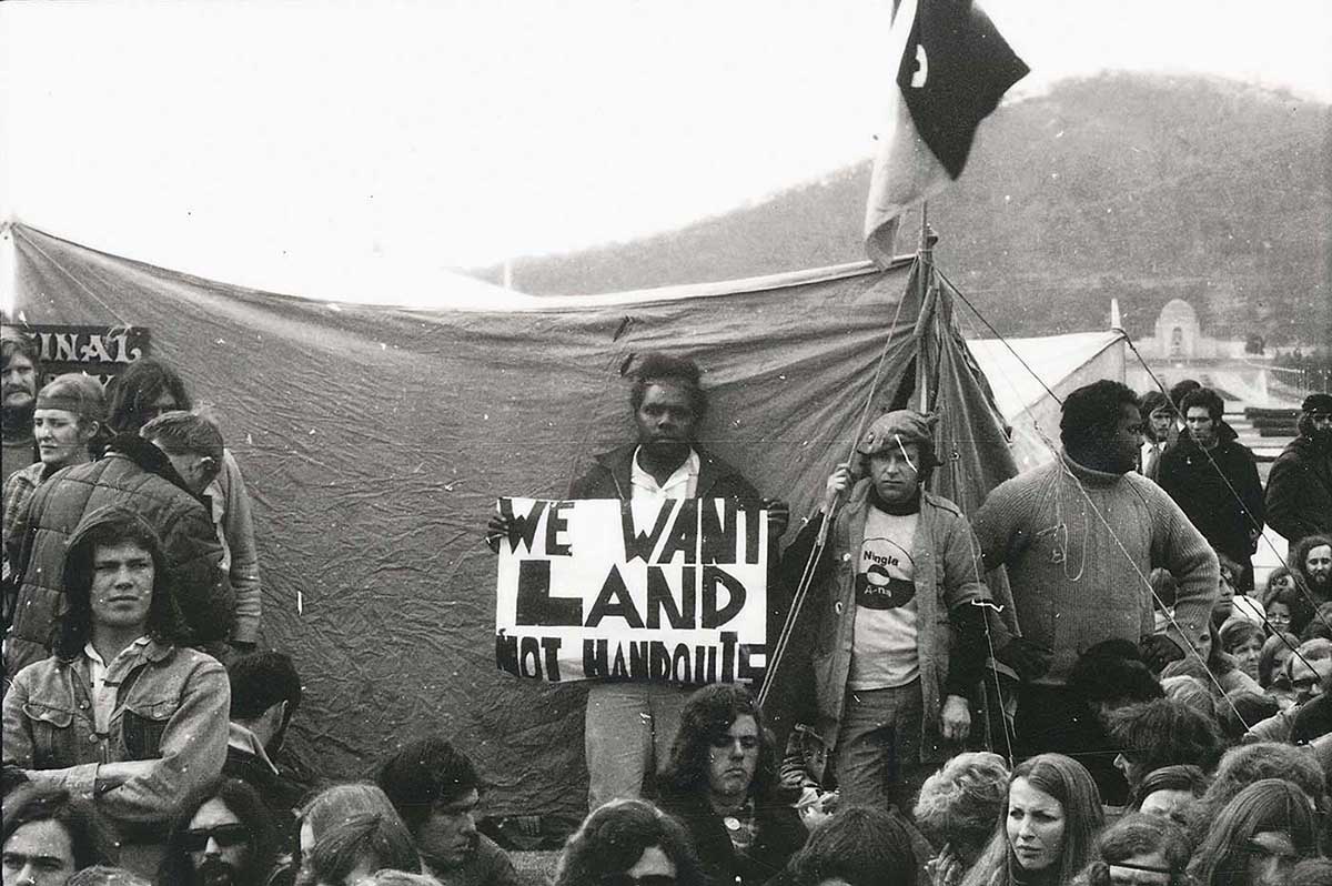 Group of young largely people, Indigenous and non-Indigenous, sitting or standing in front of a tent with a flag above it. A man at the centre of the photo stands, looking at the camera, holding a placard that says ‘We want land not handouts’. The war memorial is visible in the background.