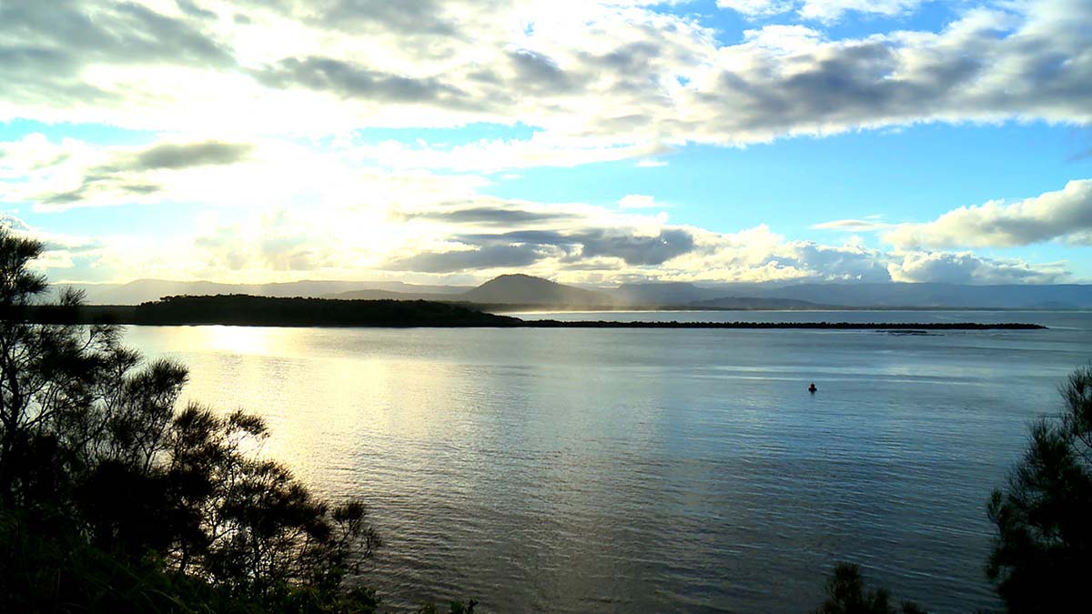 A photo of a coastal shoreline. The sky is blue with fluffy greyish clouds; sun on the far left. To the centre right is the silhouette image of a person in the water.