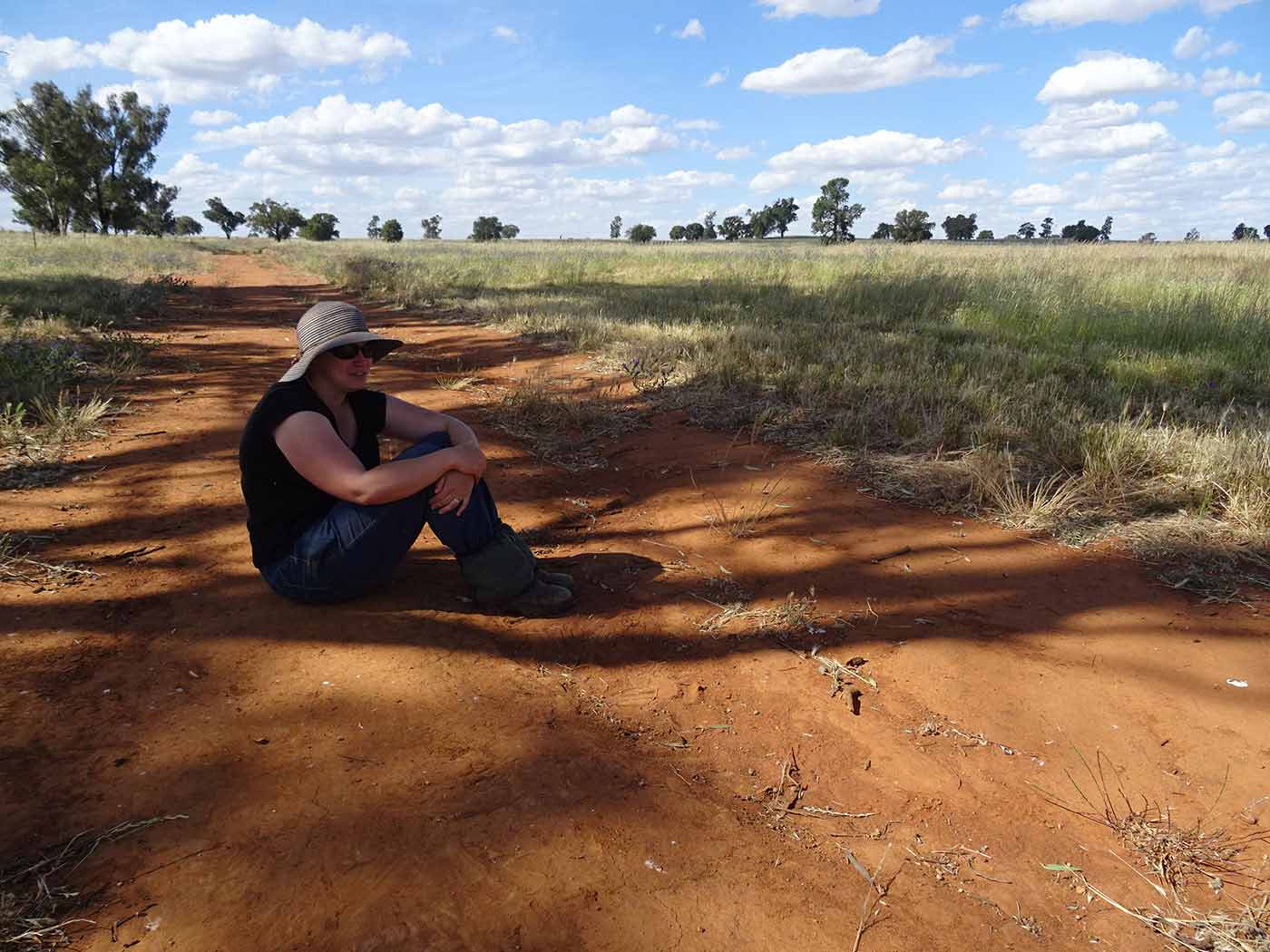 A woman sits on a path of red earth, in a paddock.