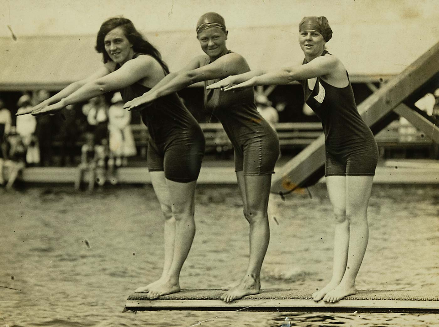 A black and white photo of Fanny Durack, Mina Wylie and Jennie Fletcher dressed in swimming costumes and standing with arms outstretched on a diving board above a swimming pool.