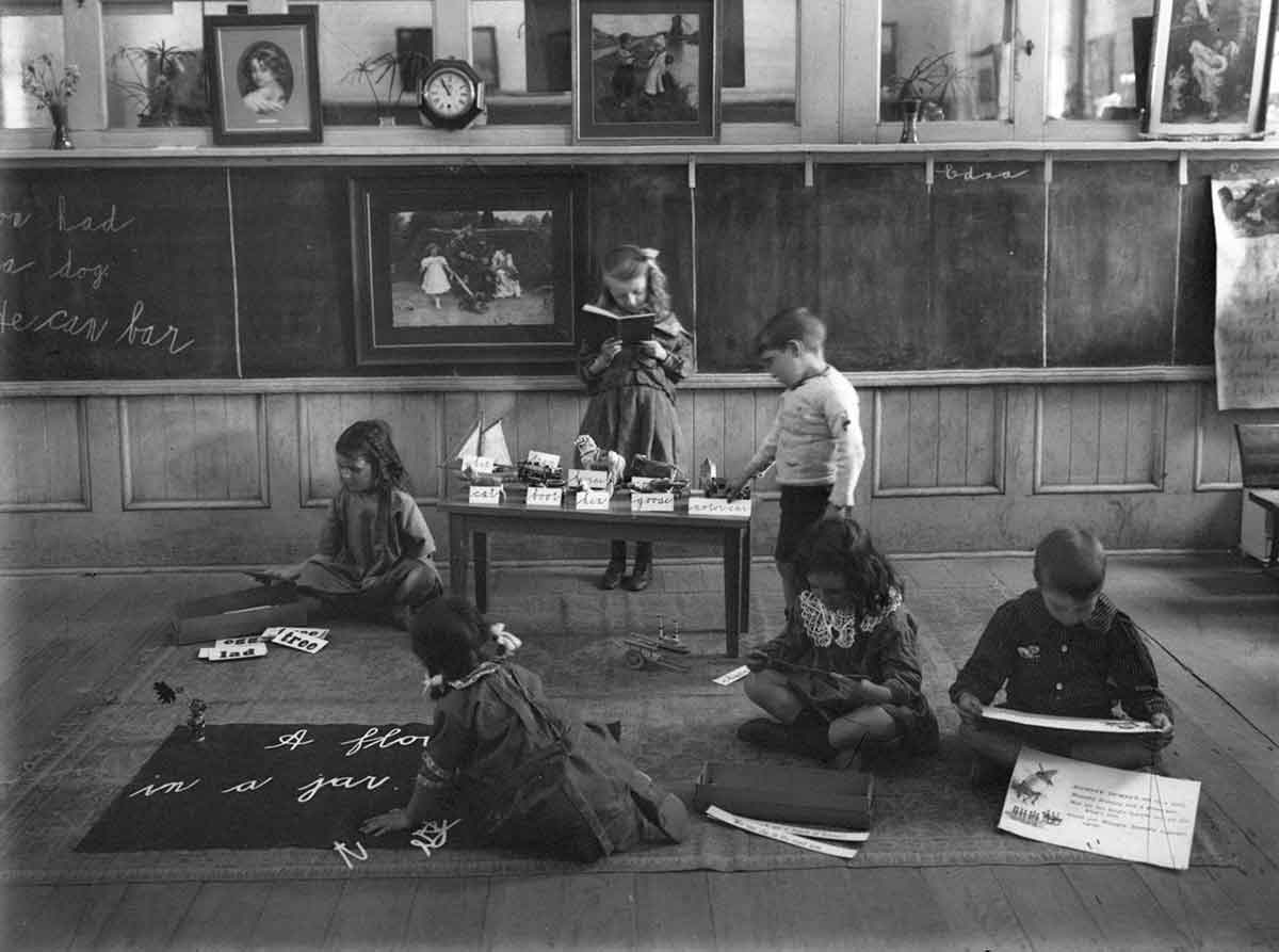 Black and white photograph of six children in a classroom. There are no desks in the classroom, with the children sitting and standing as they read and play.