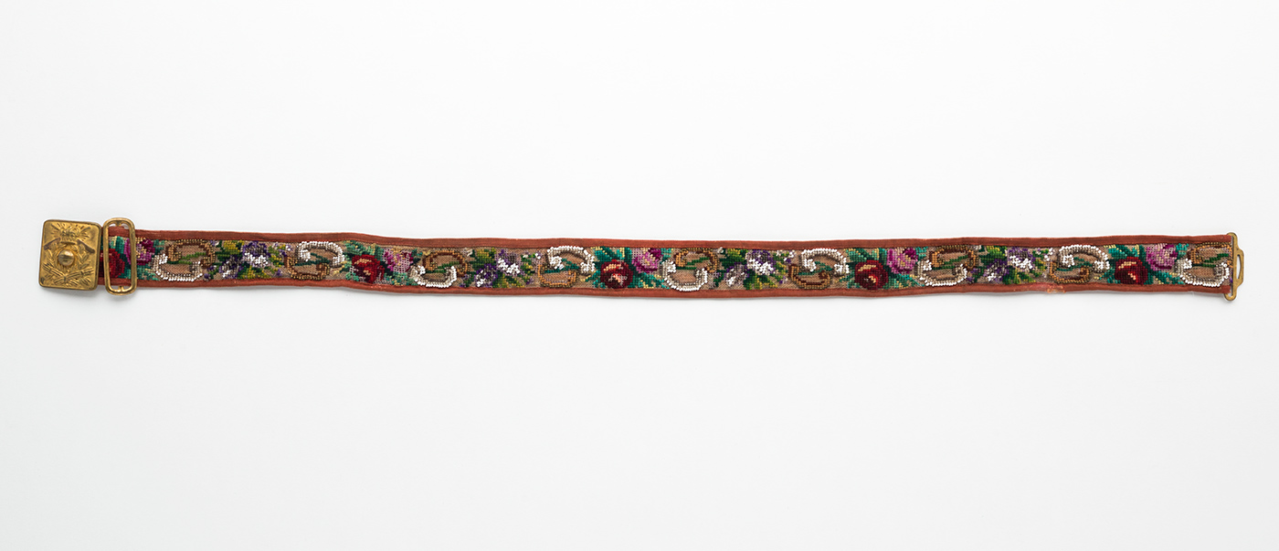 Belt of rust-coloured grosgrain backing and border, with central front strip of loose weave linen canvas gauze decorated with cross-stitch and beading with glass, haemitite and malachite beads. Pressed metal belt buckle with embossed crest consisting of crown bearing Maltese cross with two cricket bats crossed, a cricket ball in the centre and two sets of cricket stumps beneath. Metal sliding adjuster next to this and buckle hook at opposite end. Maker's stamp on buckle, consisting of a diamond shape divided into five sections containing