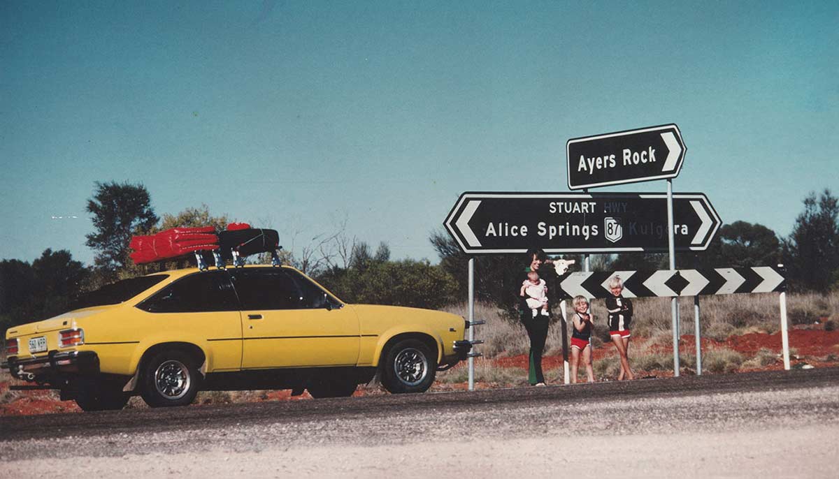 Colour photo showing a woman and three children in front of a road junction, with signs pointing to 'Ayers Rock' and 'Alice Springs'. Beside them is a bright yellow car.